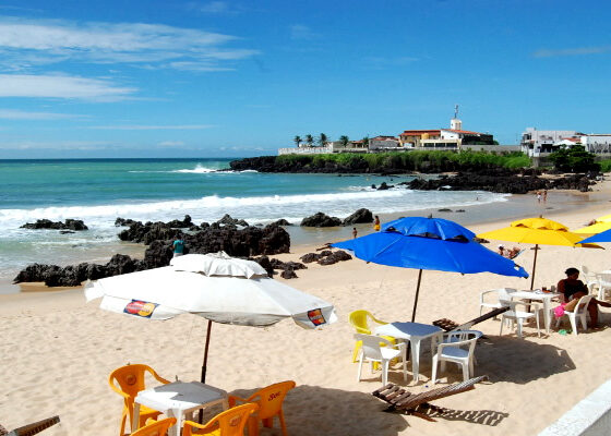 na imagem, vemos um mar, areia e algumas barracasd de praia nas cores azul, amarelo e branco. s foto é um registro da Praia dos Artistas.